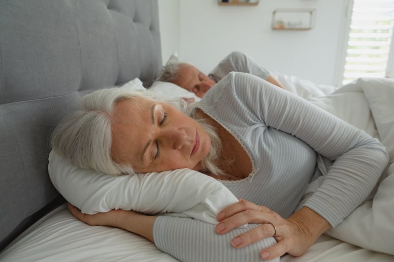 3 Health Benefits of Using Your AC While Sleeping. Photo of an elderly couple sleeping soundly in their bed.